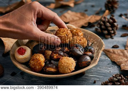 closeup of a man taking a panellet, a typical confection of Catalonia, Spain, eaten traditionally in All Saints Day, from a plate with some more and a roasted sweet potato and some roasted chestnuts