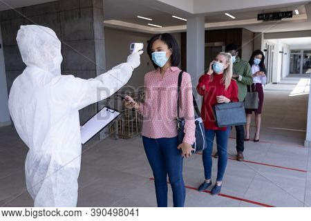 Multi ethnic group of colleagues having temperature taken by worker in protective suit before being allowed in to work. Hygiene and social distancing in workplace during Coronavirus Covid 19 pandemic.