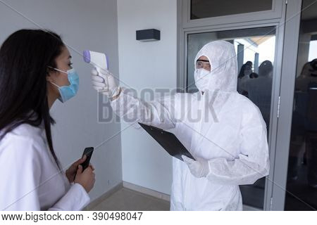 Mixed race businesswoman having temperature taken by worker in protective suit before being allowed in to office. Hygiene and social distancing in workplace during Coronavirus Covid 19 pandemic.
