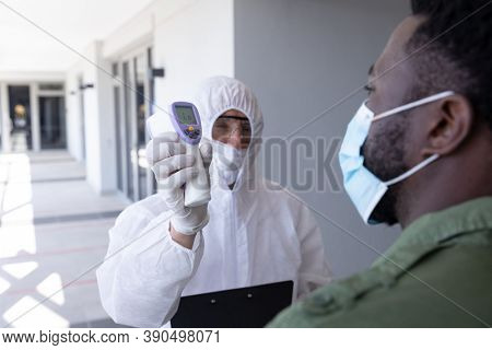 African American businessman is shown his temperature by worker in protective suit before being allowed in to office. Hygiene and social distancing in workplace during Coronavirus Covid 19 pandemic.