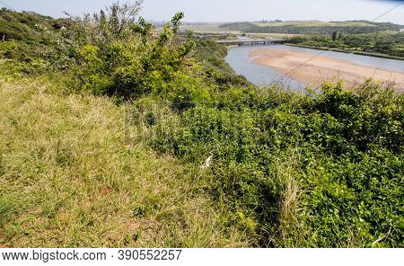 River Bordered By Natural Bush And Vegetaton With Bridge In Background