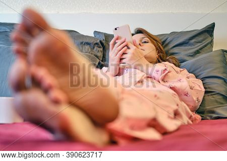 A Woman With A Phone During Quarantine Due To Coronavirus. A Red Haired Woman In Red Pajamas Is Lyin