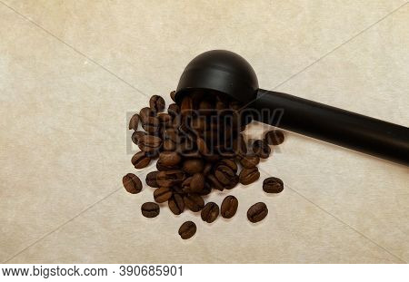 Coffee Beans Pouring Out Of A Black Plastic Spoon Against A Parchment Background.