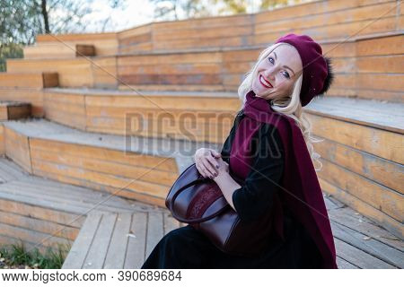 A Joyful Lady Sits On A Summer Theater Bench, Made Of Wood In A Burgundy Coat And Biret, An Adult, I