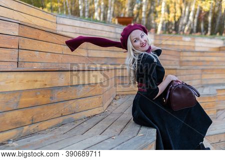 A Beautiful Lady Sits On A Wooden Summer Theater Bench In A Burgundy Coat And Biret, Looking At The 
