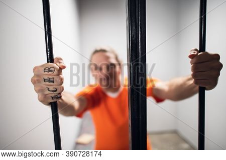 Harsh Wild Prisoner Criminal Maniac With Tattoos Grabs The Bars. A Close-up Portrait In A Prison Cel