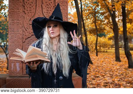 A Witch In A Hat Reads A Spell From A Book. Halloween, A Young Woman At The Carnival In The Guise Of