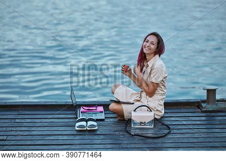 Smiling Happy Girl Holding Cup Of Coffee. Sweet Female With Laptop Near Water. Distance Learning And