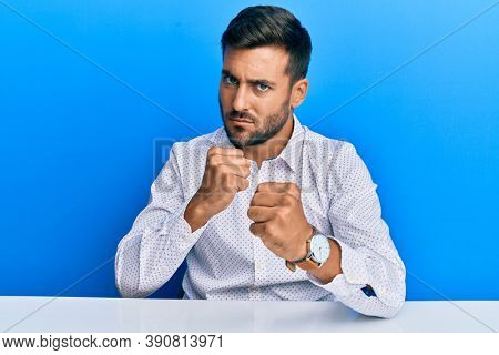 Handsome hispanic man wearing business clothes sitting on the table ready to fight with fist defense gesture, angry and upset face, afraid of problem 