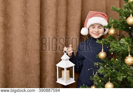 A Child With His Eyes Closed In A Striped Cap, Stands And Smiling Behind A Christmas Tree With A Lan