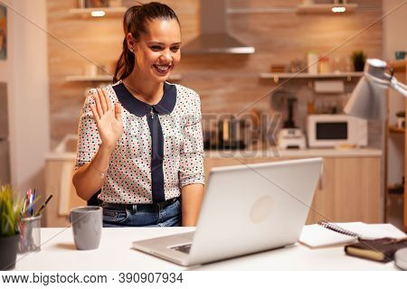 Cheerful Woman Waving During A Video Call While Working Late At Night From Home Kitchen. Employee Us