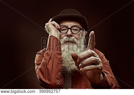 A wise old man with a long white beard in a bowler hat and glasses gives advice and shows his index finger. Black background. 
