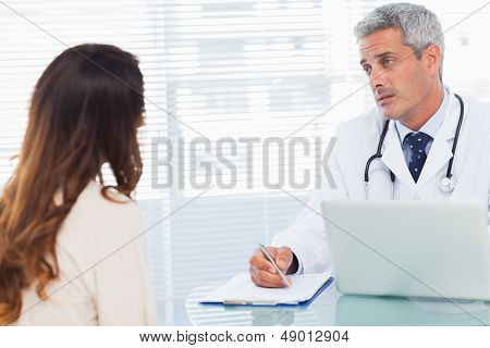 Vážné lékař mluvil se svým pacientem a psaní na notebooku v ordinaci