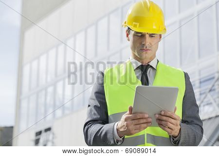 Young male engineer in reflector-vest and hardhat using digital tablet outside industry