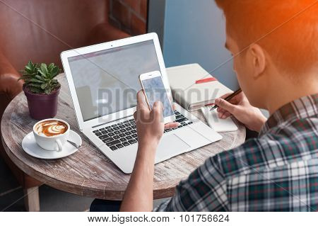 Businessman Using Smartphone And Laptop Writing On Tablet On Wooden Table In Coffee Shop With A Cup