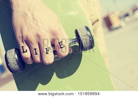 closeup of a young man with the word life tattooed in his knuckles holding a skateboard