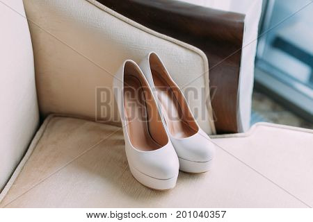 Elegant Stylish White Wedding Shoes On Wooden Table With A Dress In Background