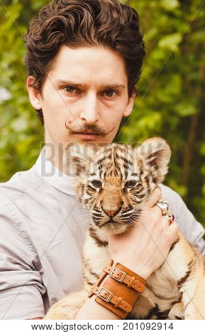 A man with a mustache looks into the face of a tiger cub otdoors