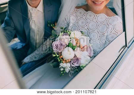 Beautiful bride in a white dress and groom in tuxedo are sitting in a car and holding a wedding bouquet. Groom embrace woman by the waist. Outdoors. Artwork