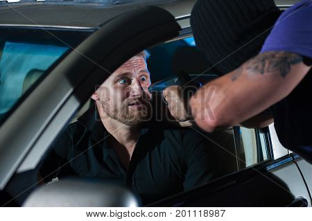 Closeup of dangerous criminal man pointing gun at the driver. Car theif threatening frightened driver, night time.