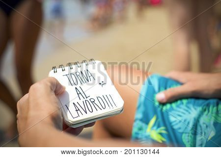 closeup of a young man sitting in a deck chair in the seashore of a beach showing a spiral notepad with the text torna al lavoro, back to work in Italian, handwritten in it