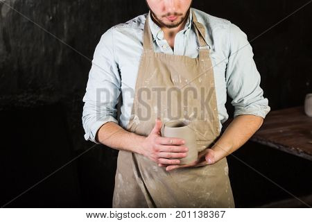 pottery, stoneware, ceramics art concept - young man in a dirty apron stand at a workshop, master hands holds cup of unfired clay, male examines a fresh product before further processing, front view