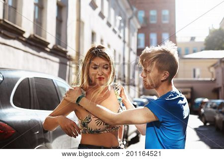 Please don't leave me. Unhappy remorseful young bearded man standing on city street holding hands on his girlfriend's shoulders asking her to forgive him. Harassment quarrel fight and disagreement