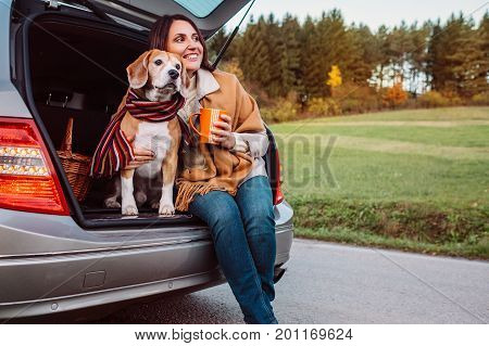 Woman and dog with shawls sits together in car trunk on autumn road