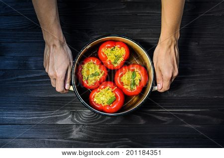 Chef holding a pan with four red stuffed peppers on dark rustic kitchen table background top view. Cooking process