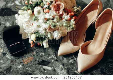 Luxury wedding rings in black box near beautiful white floral wedding bouquet and beige high heel shoes bridal accessories closeup