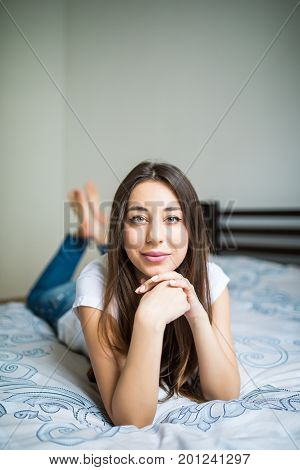 Young Woman Lying At The End Of The Bed Smiling, With Her Head Resting Upon Her Hand With The Other