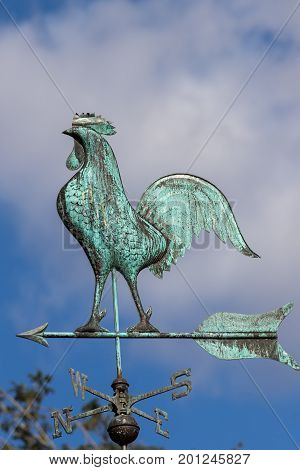 Big cockerel on a weathervane. Metal weather pointing north west. Traditional blue green windvane. Weathercock against sky.