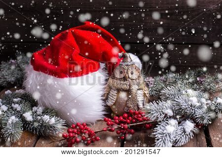 Christmas holiday composition with place for text. Santa hat decorative owl branches fur tree and red berries on aged wooden background. Selective focus. Drawn snow effect.