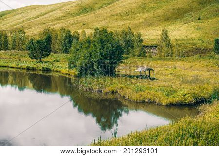 Abandoned summer alcove near beautiful pond with calm water next to meadow with native grasses trees and hills in distance; Altai mountains Russia Aya district