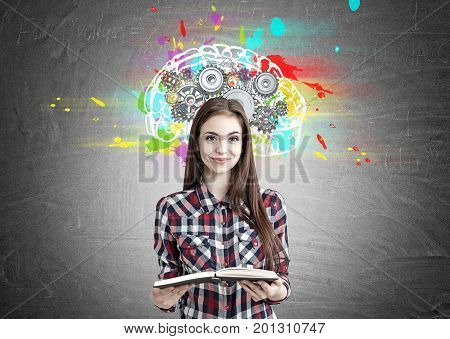 Portrait of a smiling beautiful teenage girl in a checkered shirt holding an open book and standing near a blackboard with a colorful brain sketch and gears on top of it