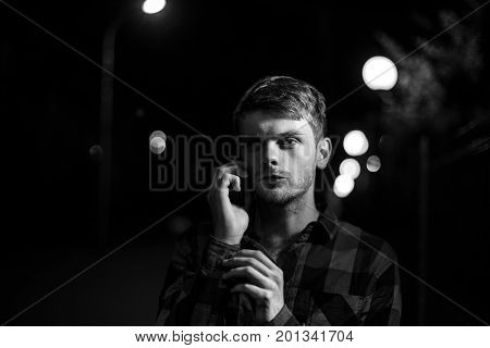 Night portrait of a hipster man. Dramatic light. He wears a plaid shirt and talks on his cell phone. Lights of the night city in the background. Black and white photography