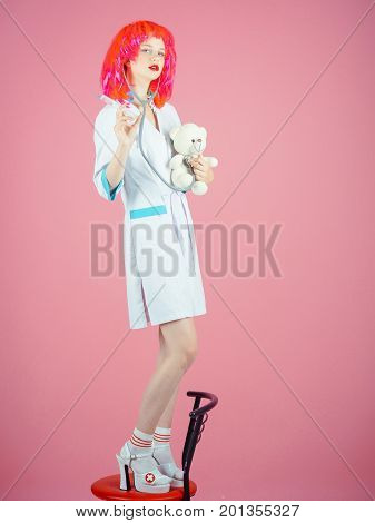 Nurse standing on chair on pink background. Girl examining teddy bear toy with syringe and stethoscope. Doctor and patient. Woman wearing medical uniform and red wig. Health care and cure concept.