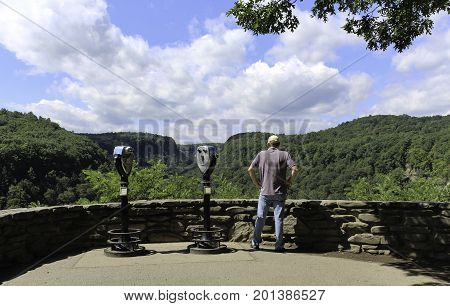 Man viewing the forest and blue skies with white clouds. Coin operated binoculars in a stone walled overlook at Letchworth State park in New York.