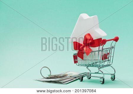 Small shopping cart with white house model decorated red bow ribbon dollars money and bunch of keys on green background. Buying a new home gift or sale of real estate concept.