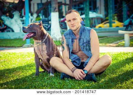 Happy friends man and dog american pit bull terrier sitting on grass in park. Caucasian male with strong dog