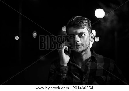 Night portrait of a hipster man. Dramatic light. He wears a plaid shirt and talks on his cell phone. Night city lights in the background. Black and white photography