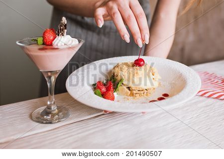 Process of decoration sweet desserts by culinary specialist in restaurant. Final touch on delicious napoleon cake with cherry and ready made fruit souffle nearby, free space below