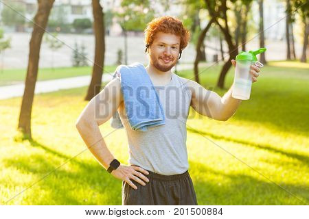 Funny redhead muscular man showing bottle and lookingat camera. Outdoor shot