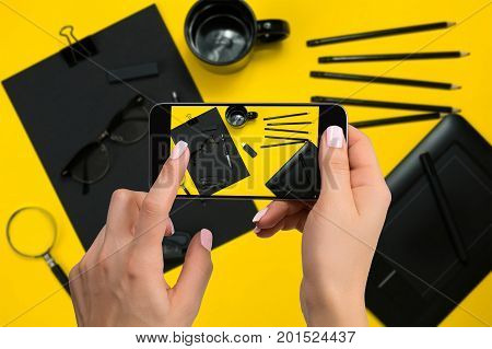 Shooting black stationery on phone's camera. Stationery on yellow background. Close-up woman's hand holding phone.