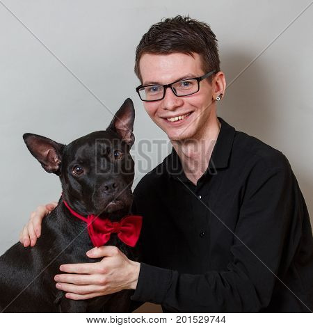 The young smiling man in glasses and black shirt holds black american staffordshire terrier dog in red bow tie looking at camera. Portrait is in studio on a grey background