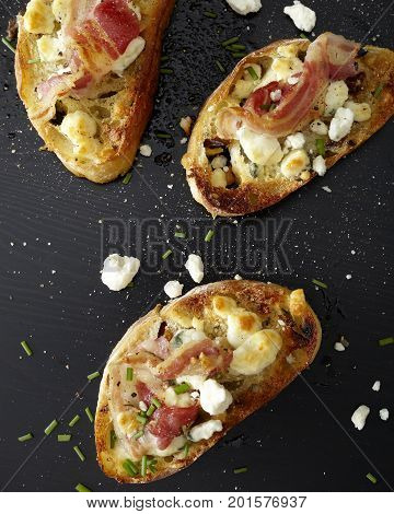 Three toast crustinis with prosciutto, herbs and cheese on a black wood surface