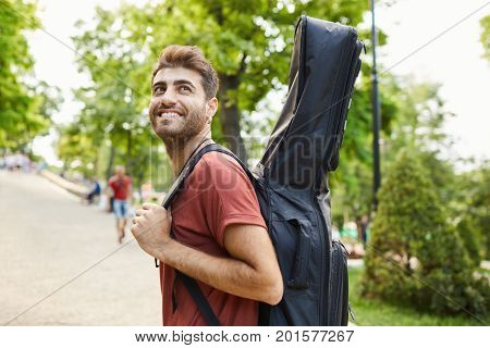 Good-looking smiling street musician walking in the street with guitar in case. Stylish happy guy carrying his guitar in case, enjoying summer day and walk.