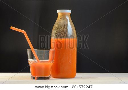 A bottle of carrot juice and a glass of carrot juice with a straw on white wood table on black background. Still life of a healthy eating concept