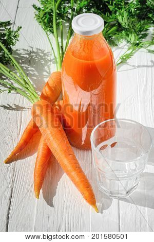 A bottle of carrot juice with empty glass and some fresh carrots on white wood table. Still life of a healthy eating concept