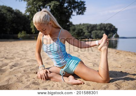 Blonde long haired girl in a blue suit is stretching and doing yoga on a beach in sunlight of the rising sun